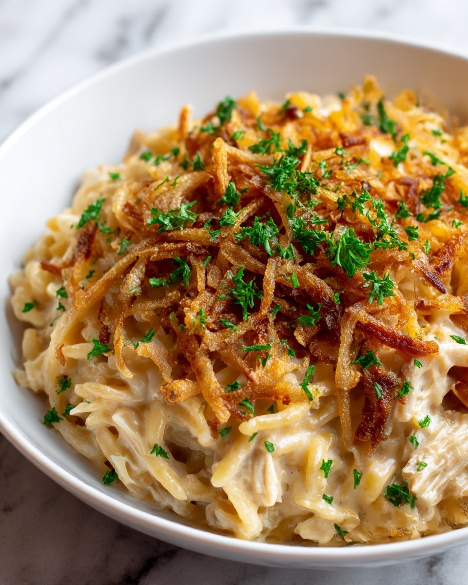 A white bowl filled with a creamy pasta dish showing two main layers: the bottom layer is a soft mixture of light beige noodles with visible cream sauce and pieces of chicken mixed in, while the top layer is golden brown crispy fried onions scattered generously with small bright green parsley leaves sprinkled all over, adding fresh color contrast. The creamy texture of the pasta and the crunchy texture of the onions are clearly visible. The bowl rests on a white marbled surface. photo taken with an iphone --ar 4:5 --v 7