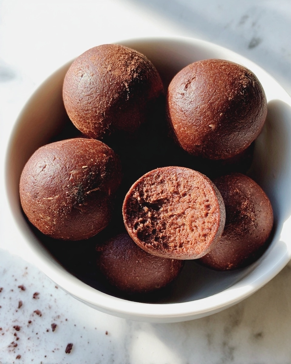 A white bowl filled with seven round, smooth, dark brown balls that have a slightly cracked surface texture. One ball is bitten, showing a dense, moist inside with a rich brown color. The bowl sits on a white marbled surface, with tiny dark crumbs scattered nearby. The lighting is natural, highlighting the soft texture of the balls. photo taken with an iphone --ar 4:5 --v 7