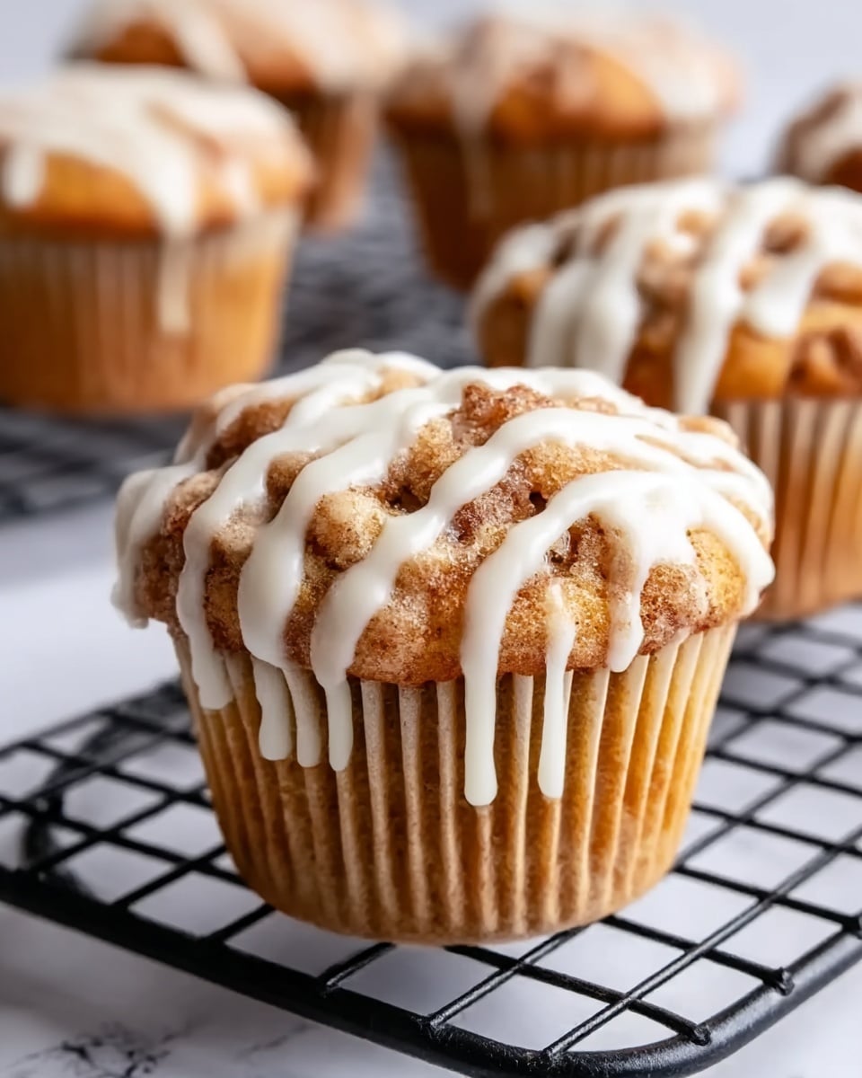 A close-up of a single muffin placed on a black round wire cooling rack, surrounded by several muffins blurred in the background. The muffin has a golden-brown color with a rough, crumbly texture on top, covered with a drizzle of thick, white icing that runs down slightly over the edges. The muffin liner is light brown with thin darker stripes. The photo is set on a white marbled surface. photo taken with an iphone --ar 4:5 --v 7