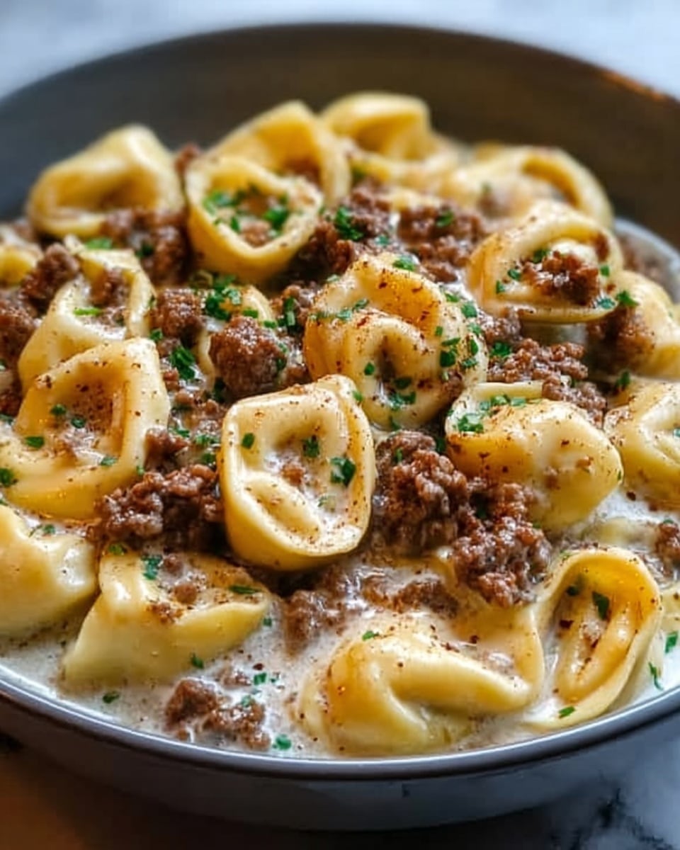 A close-up of a black bowl filled with tortellini pasta covered in a creamy sauce, topped with browned ground meat scattered evenly throughout, and garnished with small green herb pieces. The tortellini are a light yellow color with a smooth texture, and the sauce looks thick and rich, pooling slightly at the bottom of the bowl. The bowl sits on a white marbled surface, and the lighting highlights the glossy shine on the sauce and the pasta folds. Photo taken with an iphone --ar 4:5 --v 7