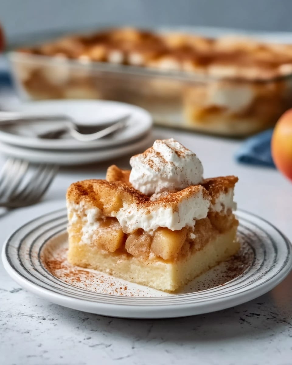 A square piece of apple cinnamon dessert with three visible layers sits on a white plate with subtle grey rings. The bottom layer is a soft, pale golden crust, followed by a middle layer of chunky, light brown baked apples mixed with creamy filling. The top layer shows a golden brown baked crust dusted with cinnamon and sugar, crowned with a dollop of whipped cream and a light sprinkle of cinnamon powder. Behind the plate, a white marbled texture surface can be seen with a round white plate holding silver forks and a glass baking dish full of the dessert blurred softly in the background. Photo taken with an iphone --ar 4:5 --v 7