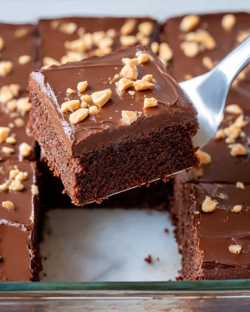 A close-up view of a chocolate dessert in a clear glass baking dish, cut into square pieces. The dessert has two visible layers: a thick, moist, dark brown cake base with a slightly crumbly texture, and a smooth, glossy dark chocolate layer on top sprinkled with small, light beige chopped nuts. One square piece is being lifted with a shiny metal spatula, showing the dense yet soft texture of the cake beneath the glossy frosting. The dessert is set on a white marbled surface. photo taken with an iphone --ar 4:5 --v 7