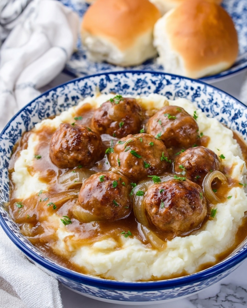 A white bowl with blue patterns holds a dish made of two main layers: the bottom layer is creamy white mashed potatoes with a soft, smooth texture, while the top layer has several round brown meatballs covered in a shiny brown gravy with visible slices of cooked onions. The meatballs are sprinkled with small green herb pieces for color contrast. In the background, two light brown soft bread rolls rest on a white marbled surface, creating a cozy setting. photo taken with an iphone --ar 4:5 --v 7