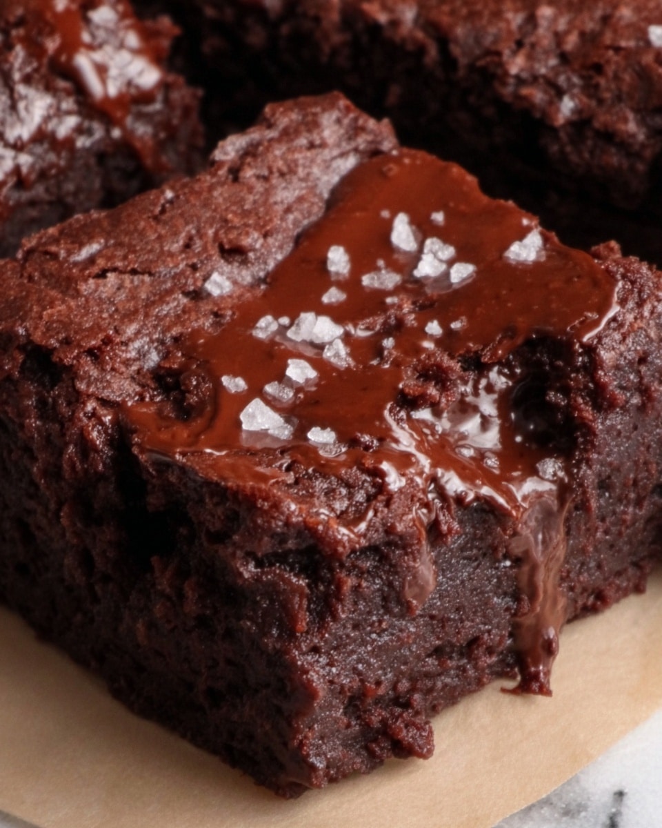 A close-up of a single square brownie with a thick, moist, and dense texture. The brownie has a deep dark brown color, with a glossy layer of melted chocolate drizzled unevenly on top. Its surface shows small cracks and a slightly crumbly texture, with a few flakes of white sea salt sprinkled on top. The brownie sits on a piece of parchment paper placed over a white marbled texture. Photo taken with an iphone --ar 4:5 --v 7