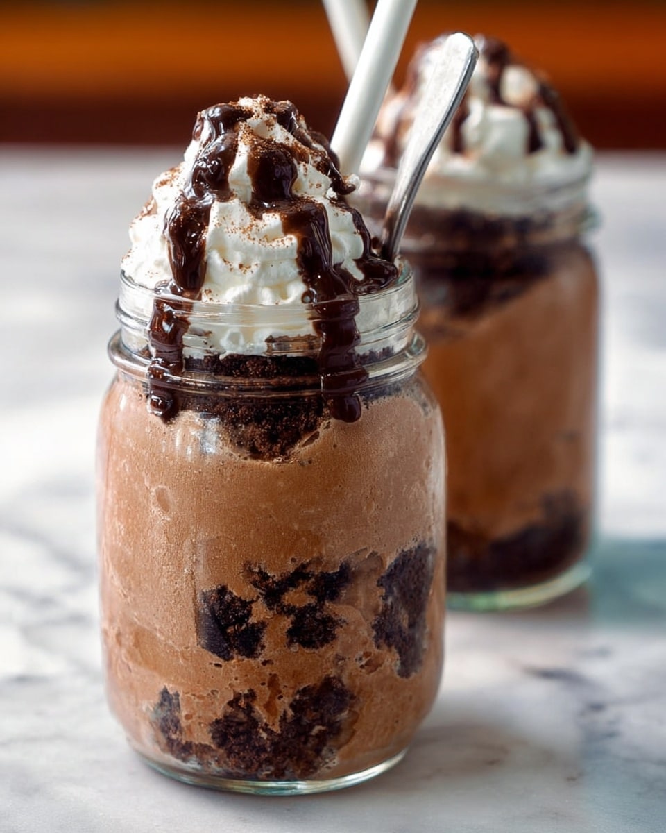 The image shows a close-up of two clear glass jars filled with a layered drink. The bottom layer is a thick, light brown chocolate mousse with visible dark chocolate crumb pieces mixed in. Above that are darker chocolate bits pressed into the mousse, creating a textured darker layer inside the jar. The top is a white whipped cream peak with a drizzle of dark chocolate syrup flowing down the jar's side. A white spoon is placed inside each jar, slightly tilted. The jars sit on a white marbled surface with a soft background blur. photo taken with an iphone --ar 4:5 --v 7