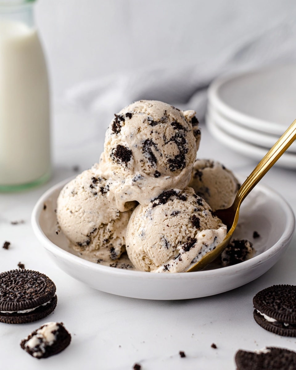 The image shows a white plate holding four scoops of cookies and cream ice cream, each scoop light beige with many dark chocolate cookie bits mixed in. The scoops are stacked closely, with a golden spoon scooping one ball on the right side. Around the plate on the white marbled surface are pieces of broken chocolate sandwich cookies with white cream inside. A glass bottle of milk is blurred in the foreground on the left, and a small stack of white plates is blurred in the background on the left side. photo taken with an iphone --ar 4:5 --v 7