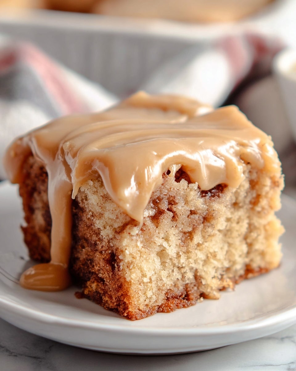 A close-up view of a thick slice of cinnamon roll cake with one visible layer, showing a soft, moist, golden-brown base mixed with swirls of dark cinnamon within. On top, there is a thick layer of smooth, light brown caramel or brown sugar frosting that drips down the sides unevenly, adding a creamy texture. The slice is placed on a glossy white plate, set on a white marbled textured surface with a soft focus background that includes a hint of a striped cloth. Photo taken with an iphone --ar 4:5 --v 7