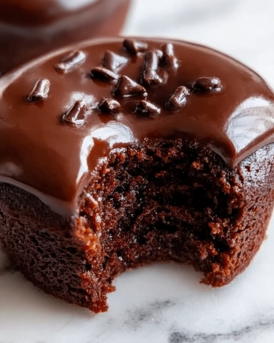 A close-up of a chocolate dessert shaped like a cupcake with a shiny, smooth chocolate glaze on top. The glaze is dark brown and glossy, with a few small shiny chocolate pieces sprinkled on the top layer. The inside is visible where a bite has been taken, showing a moist, dark brown, soft cake texture. The dessert sits on a white marbled surface. Photo taken with an iphone --ar 4:5 --v 7
