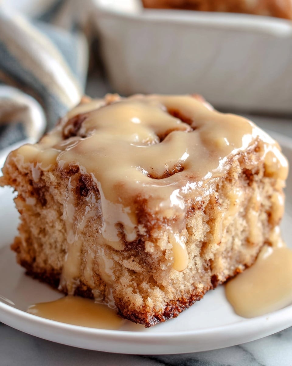 A single square piece of cinnamon roll sits on a white plate with a soft, thick layer of golden tan glaze dripping slightly down the sides. The cinnamon roll has a textured, slightly crumbly surface with visible swirls of cinnamon brown inside the light beige dough, showing a contrast between the baked outer crust and the moist inner layers. The background shows a white marbled texture and a faint glimpse of a striped cloth partially blurred behind the dish. Photo taken with an iphone --ar 4:5 --v 7