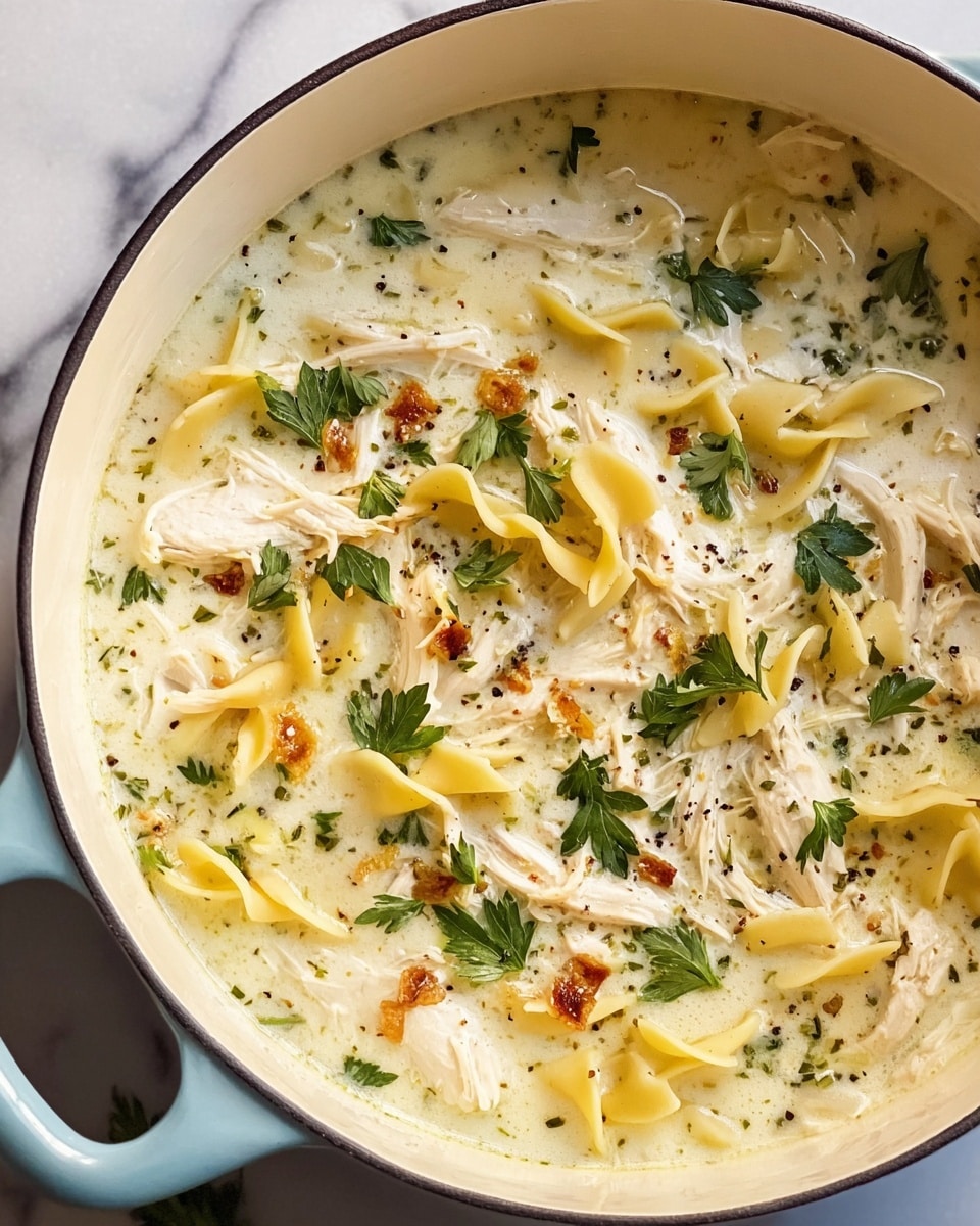 A close-up view of a pot filled with creamy soup, showing shredded white chicken pieces and small, pale yellow pasta shapes floating in a light cream-colored broth. The soup also contains scattered green parsley leaves and specks of black pepper, giving texture and contrast to the creamy surface. The pot has a pale blue outer edge with a white interior, sitting on a white marbled surface with a soft blue cloth partially visible underneath. Some fresh green herbs are placed next to the pot on the left side. Photo taken with an iphone --ar 4:5 --v 7
