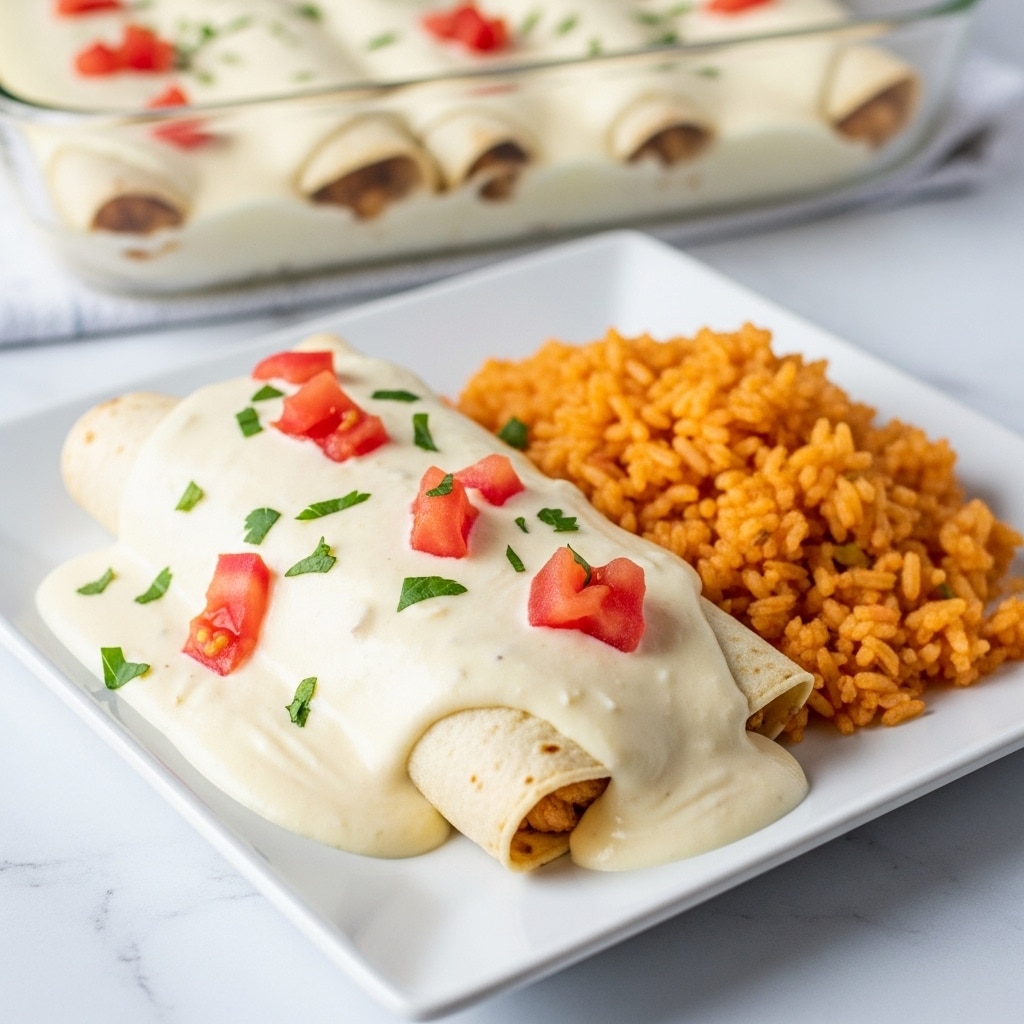 A white square plate holds a rolled enchilada covered with a thick, creamy white sauce that has visible chunks of red tomato and small green herb pieces sprinkled on top. The sauce generously drapes over the entire enchilada, partially spilling onto the plate. Next to the enchilada is a serving of reddish-orange Mexican-style rice with separated grains and a slight shine. In the background, a clear glass baking dish with more enchiladas covered in the same creamy sauce is slightly out of focus, placed on a white marbled surface. photo taken with an iphone --ar 4:5 --v 7
