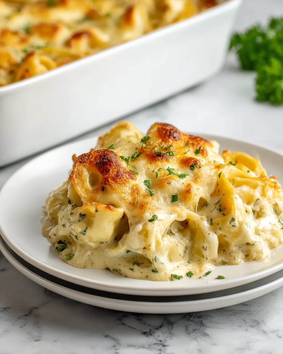 A white plate holds a serving of baked tortellini pasta covered in a creamy white sauce with melted, slightly browned cheese on top, giving it a golden crust. The tortellini are plump and round, with visible green herbs mixed into the sauce and sprinkled on top. The layers include the smooth pasta base, a thick white cheese sauce with herbs, and golden-browned melted cheese on top. In the background, a white baking dish with more of the same baked pasta is slightly out of focus, all set on a white marbled surface with a blurred cloth and green herbs nearby. Photo taken with an iphone --ar 4:5 --v 7